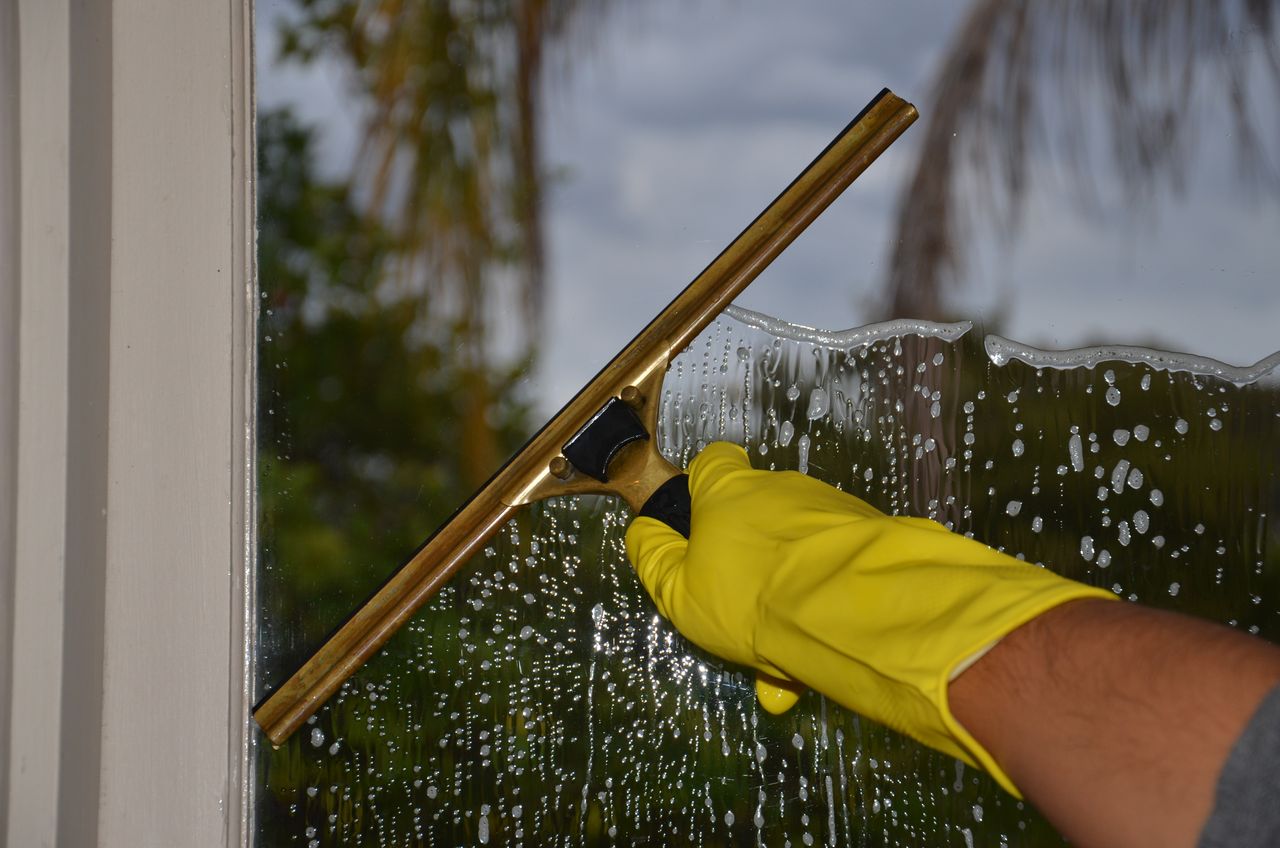 A close-up view of a hand cleaning an outdoor window with a squeegee while wearing yellow gloves.